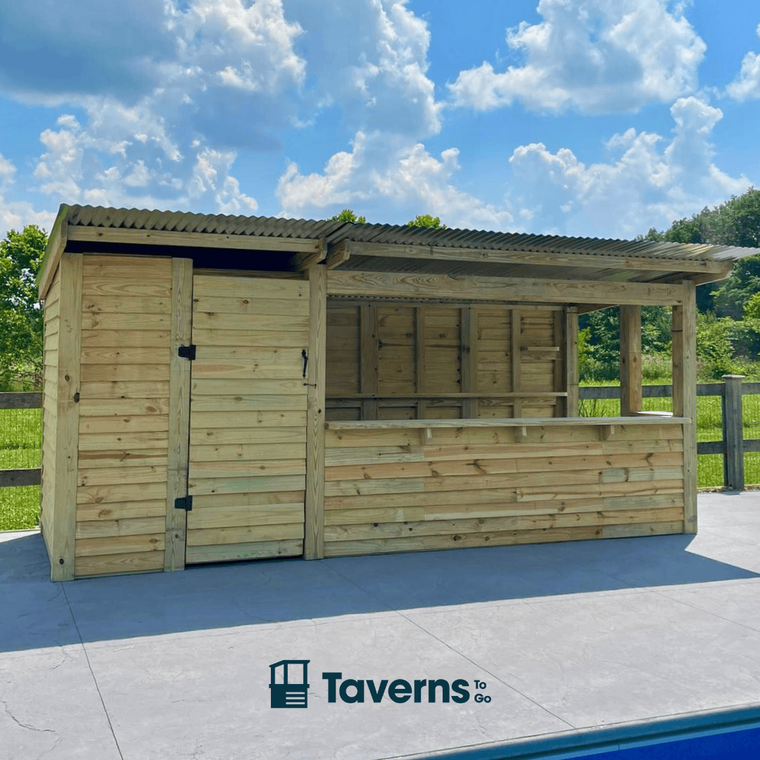 Wooden outdoor shelter with a roof, set against a blue sky with clouds and greenery.