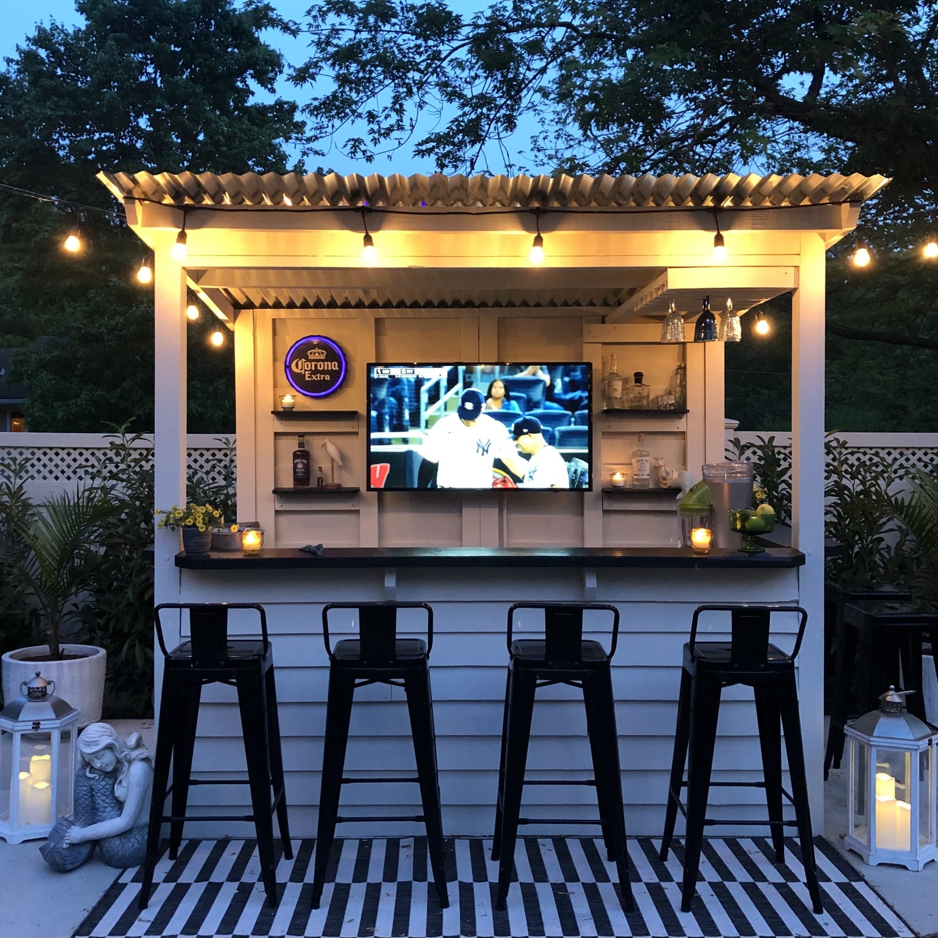 Outdoor bar area with a television screen displaying a sports event, surrounded by plants and lanterns.