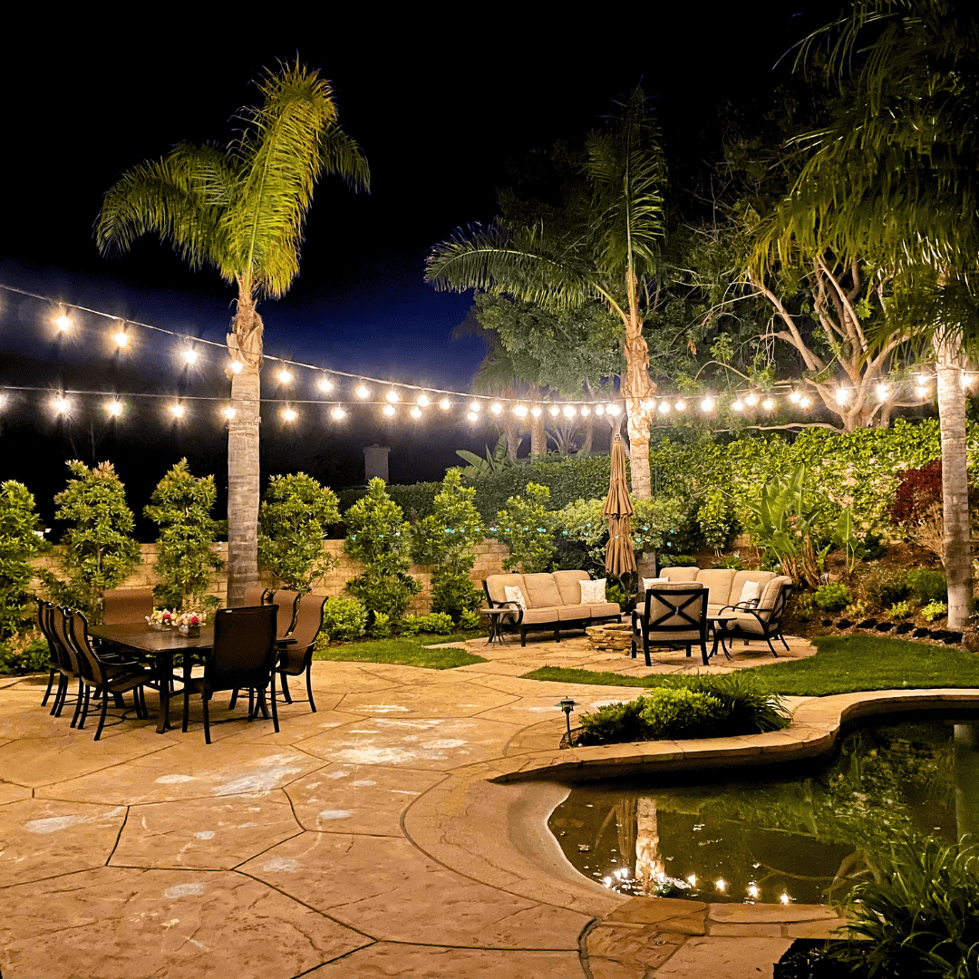 Backyard patio with string lights, outdoor furniture, and a pond at night.