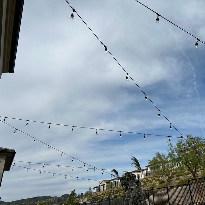 String lights hanging against a cloudy sky with houses and trees in the background.