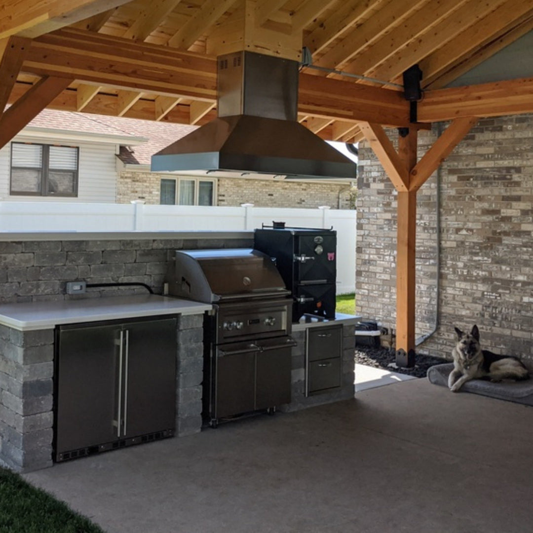 Outdoor kitchen with grill and refrigerator under a wooden pergola, dog resting nearby.